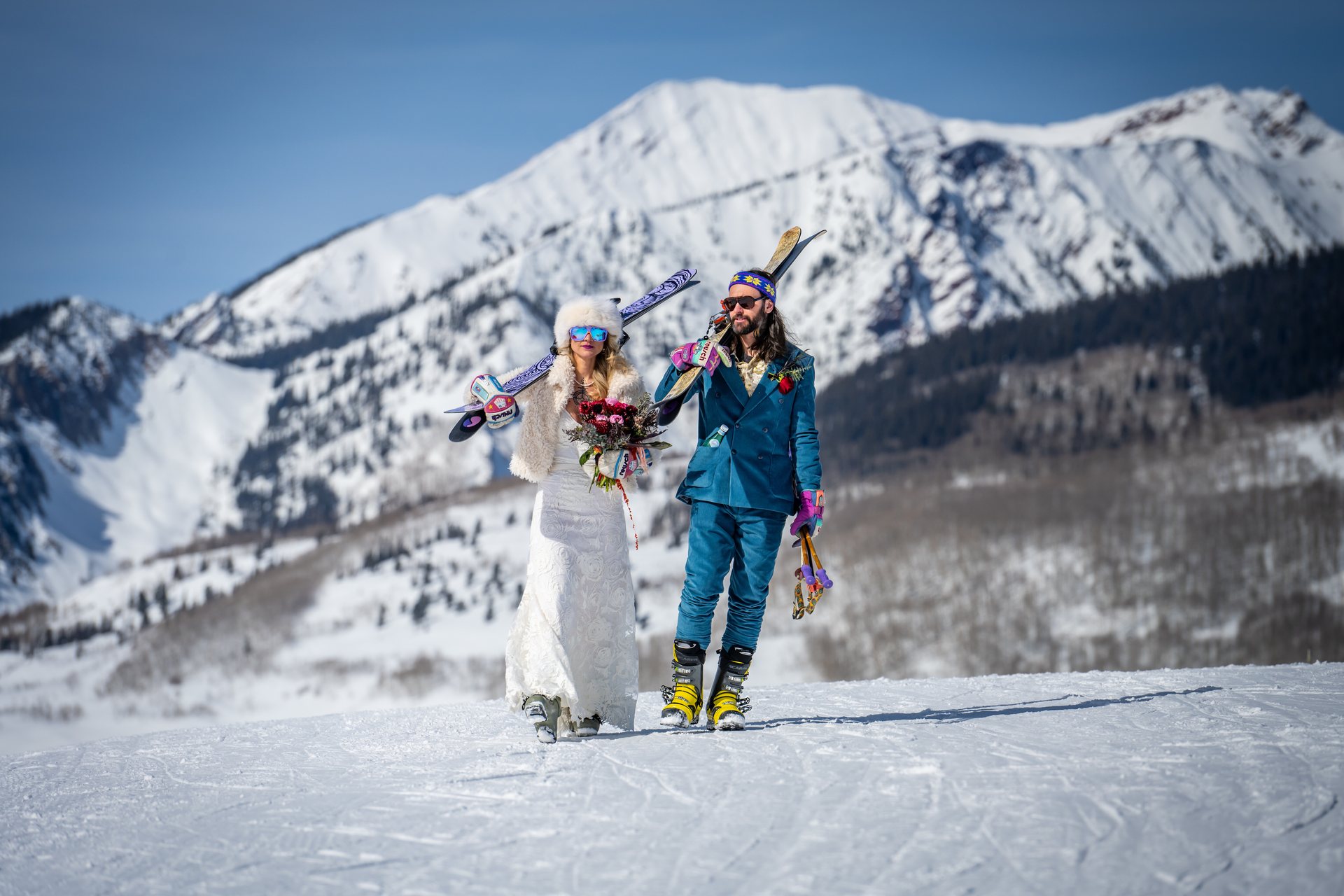 Janice and Alex in their ski-wedding attire, carrying skis below Mt. Crested Butte