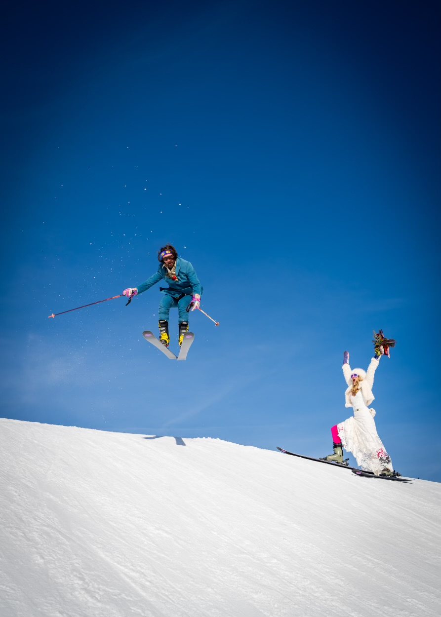 Alex catching air on skis in his wedding suit while Janice cheers below